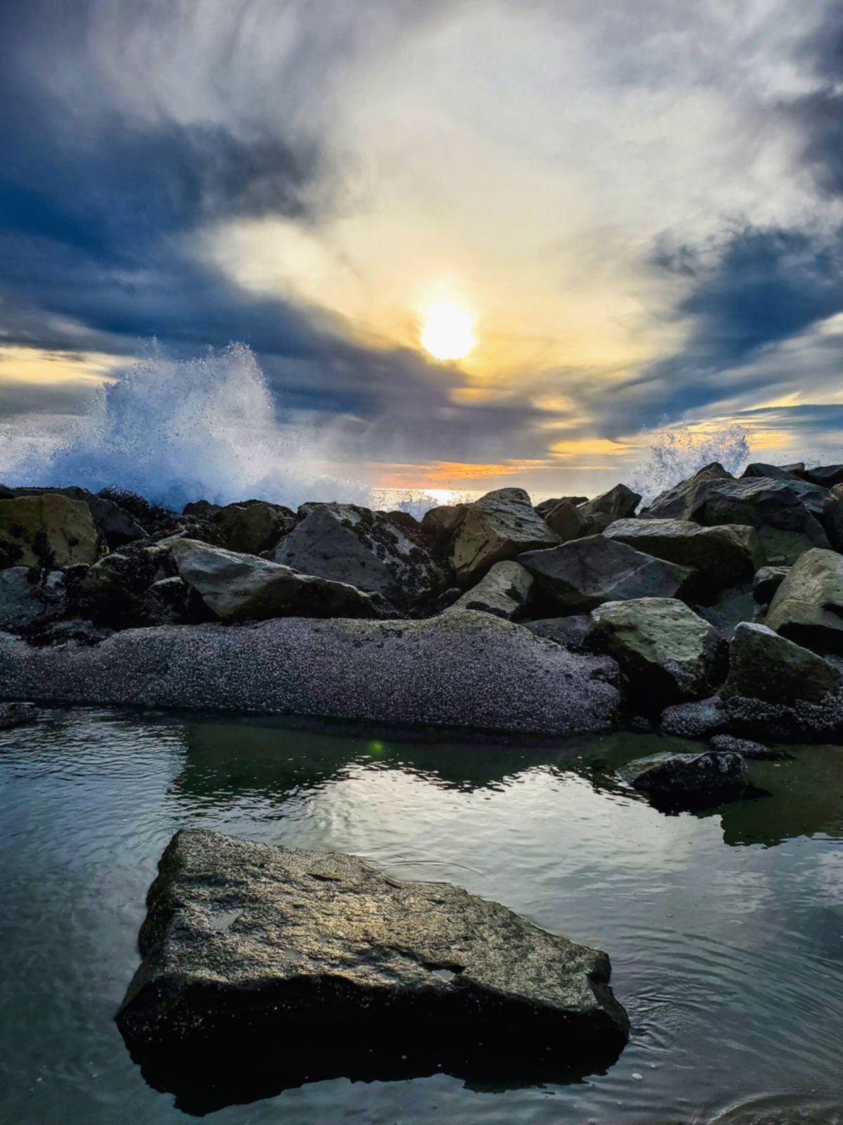 Waves at Santa Monica Beach against a rocky jetty under a cloudy sky with sun breaking through